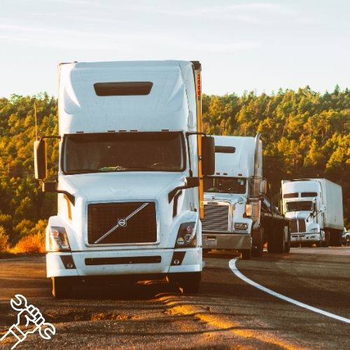 White semi-trucks in a convoy on a curved highway at sunset, with a forest backdrop; a wrench logo sits in the bottom-left corner.